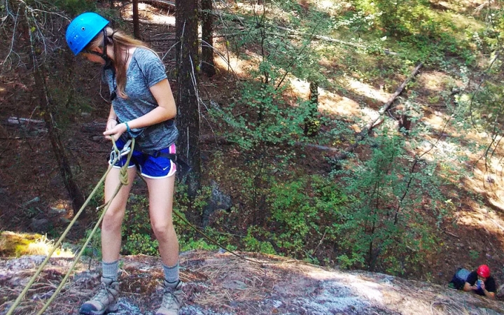 an outward bound student wearing safety gear looks down as they repel from a rock wall