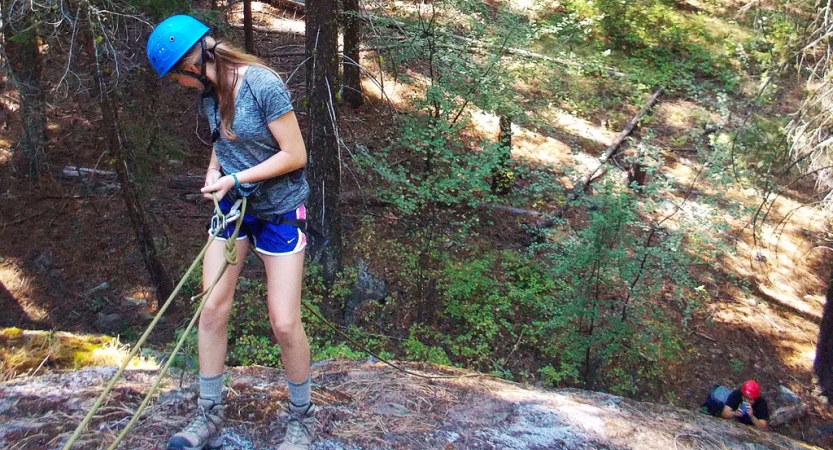 an outward bound student wearing safety gear looks down as they repel from a rock wall