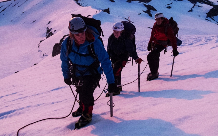 A group of outward bound students wearing mountaineering gear make their way up a snowy incline. The snow appears pink from the sunrise or sunset. 