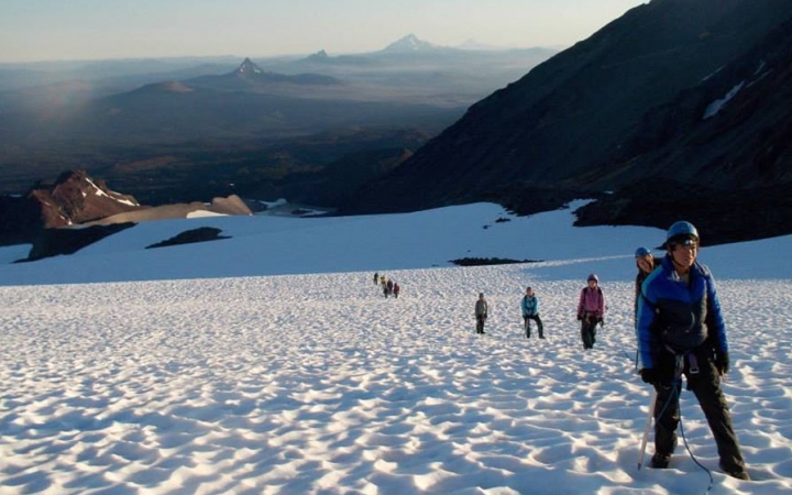 A group of outward bound students wearing mountaineering gear make their way up a snowy slope. There are mountains in the distance. 