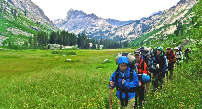 A group of young people wearing backpacks hike through a valley of bright green grass. There are mountains and evergreen trees in the background.