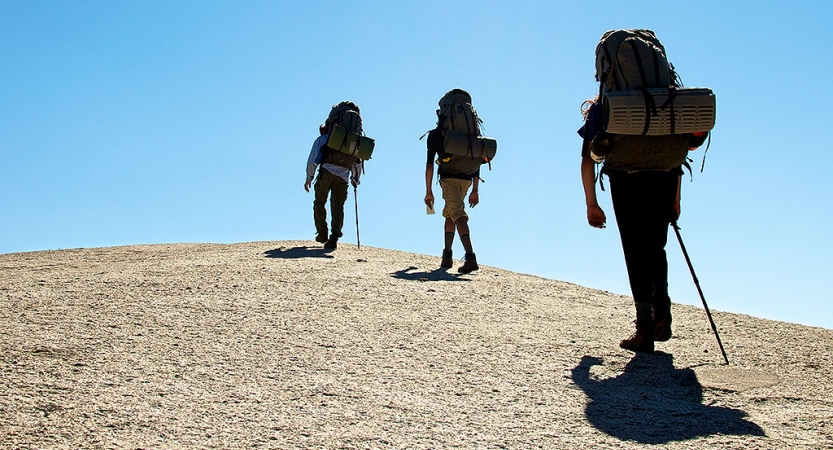 Three people wearing backpacks hike away from the camera up a rock incline. There is a blue sky above.