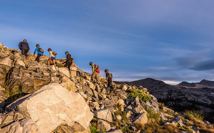 A group of outward bound students hike up a rocky incline. There are mountains in the background. 