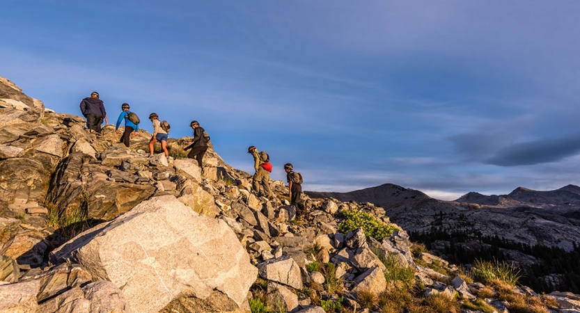 A group of outward bound students hike up a rocky incline. There are mountains in the background. 