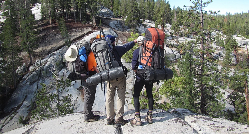 three people wearing backpacks stand on a rock while one of them points outward