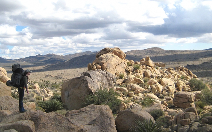 A person wearing a backpack looks out over a rocky landscape. There are mountains in the distance. 