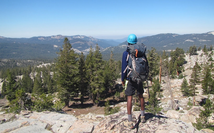 a person wearing a helmet and a backpack faces away from the camera and looks out over a vast mountainous landscape