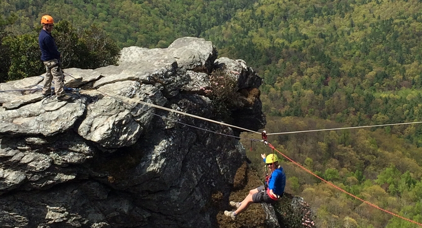 a person wearing safety gear is suspended by ropes mid air between a rock formation and another object that is not in the picture. One person stands on the rock formation, overlooking a vast wooded area. 