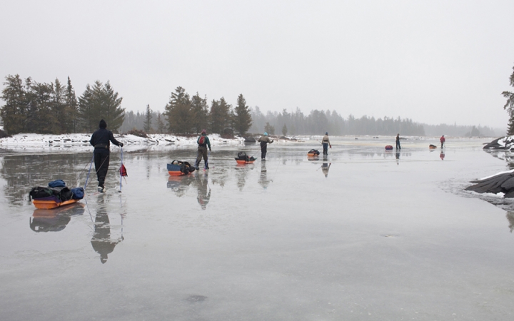 A line of people pull small sleds in a line across an icy lake