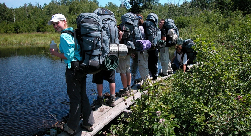 A group of people wearing backpacks stand on a narrow wooden plank over a body of water. Thick greenery surrounds them.