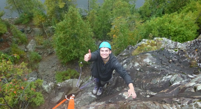 A person wearing safety gear is secured by ropes as they look up at the camera and smile while giving a thumbs up.