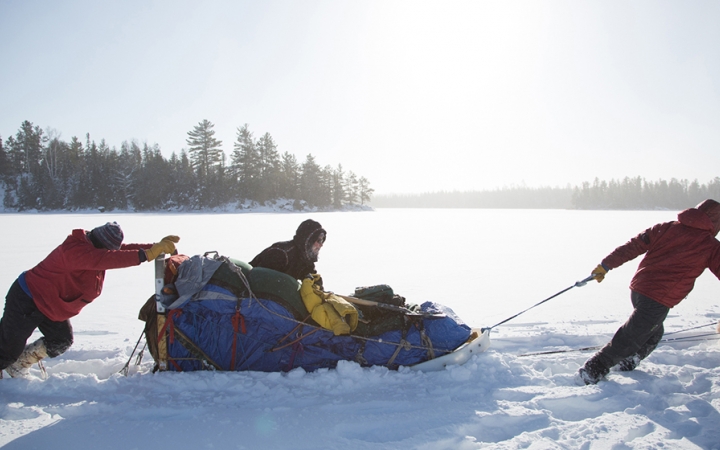 Three mushers help to push and pull a sled through thick snow. There are trees in the background. 