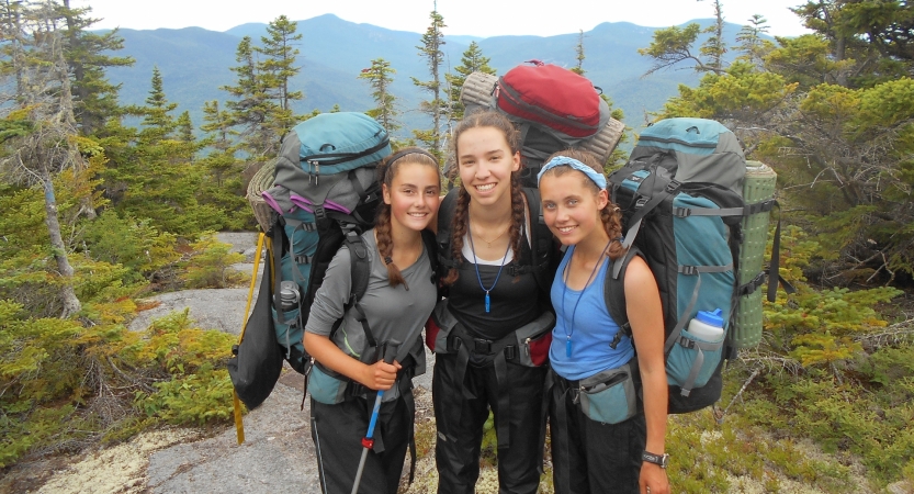 Three young people wearing backpacks smile for the photo in a wooded area with a mountain landscape in the background. 